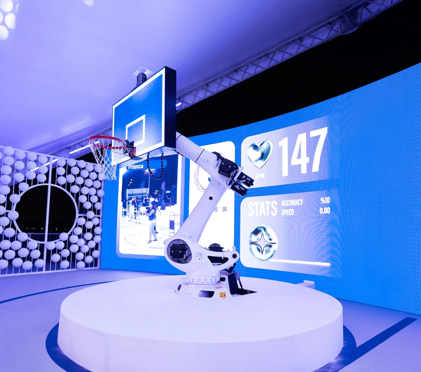 A robotic industrial arm holds a basketball hoop and backboard in a blue-lit event booth, with large screens behind showing a game interface, stats, and a score of 147.